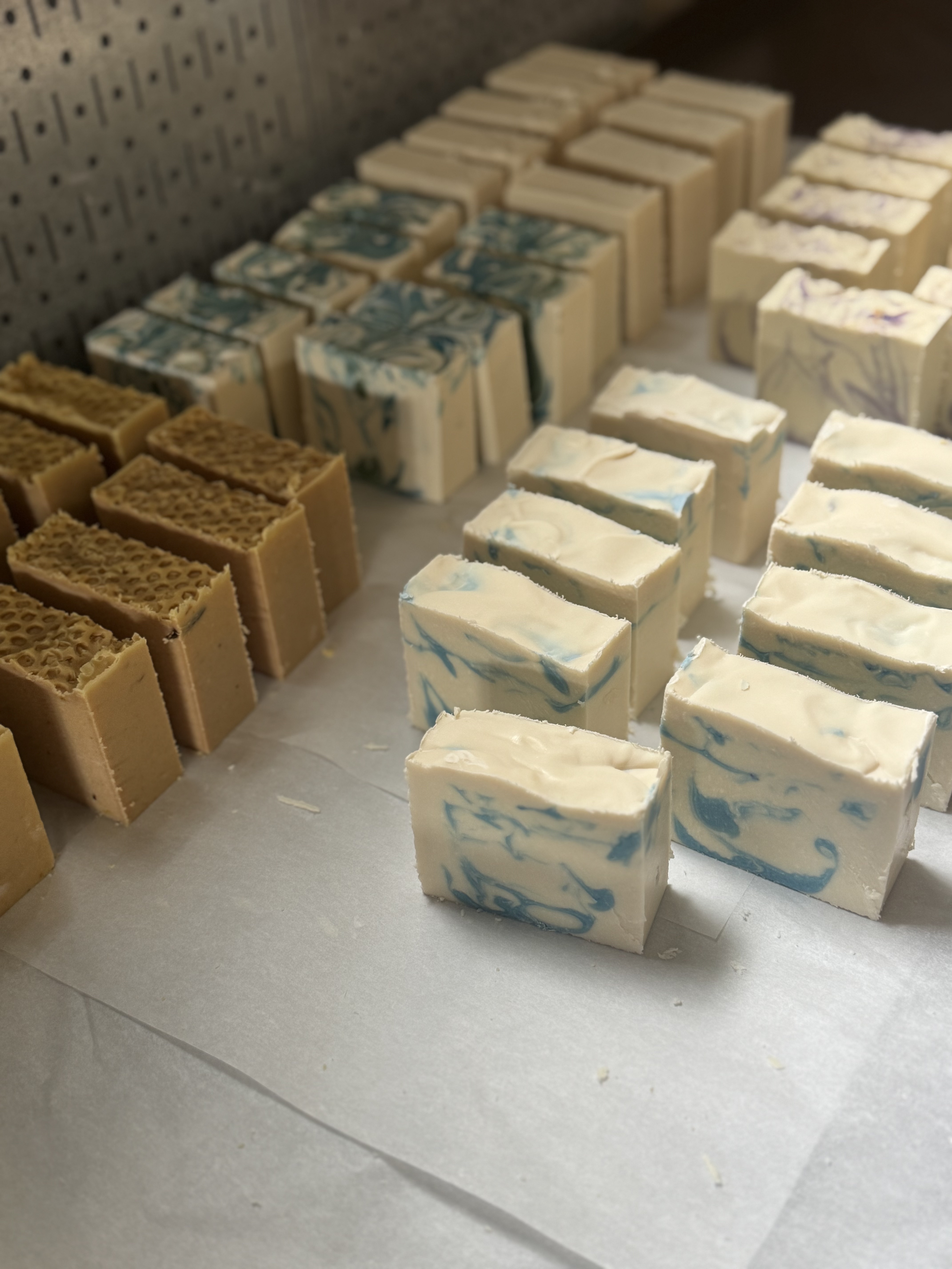 Two fresh cold process soap loaves in wooden molds side by side — a deep purple loaf with botanicals in the background and a pink swirl loaf with gold mica in the foreground.