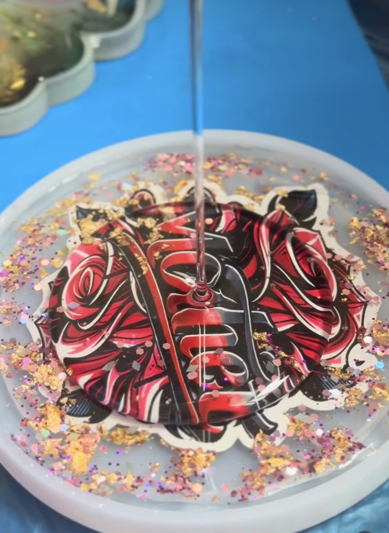 Resin epoxy coaster with bold red and black rose design, surrounded by gold and pink glitter, with clear resin being poured over it.