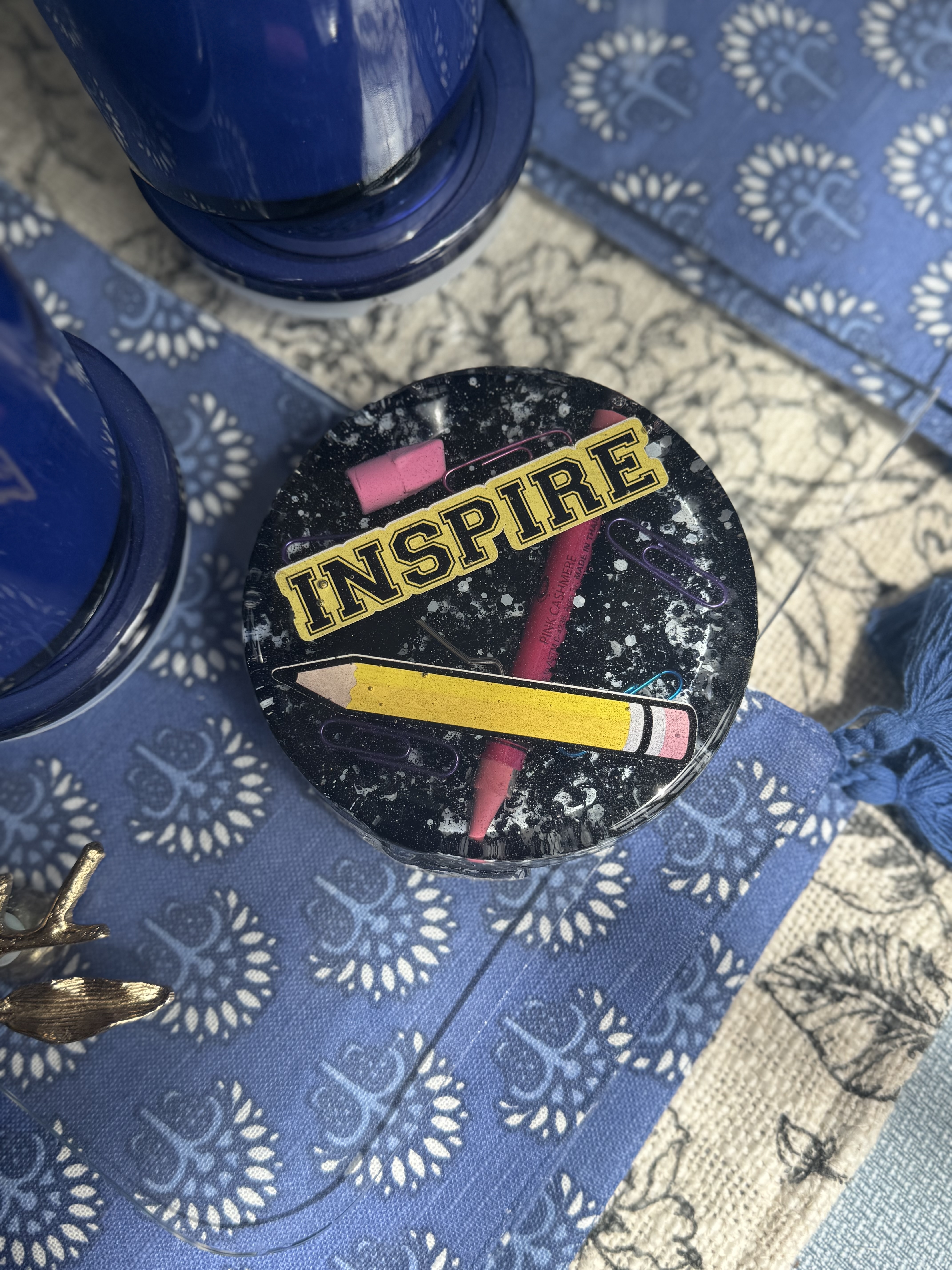 Circular resin coaster with a black, speckled background featuring a yellow pencil, pink eraser, paper clips, and the word 'INSPIRE' in bold gold letters. Set on a blue patterned fabric with a blue tumbler nearby.